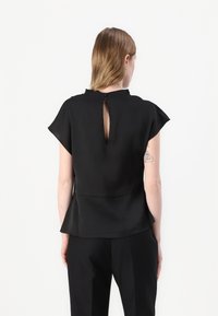 Top nero a maniche corte con dettaglio a chiave sul retro, realizzato in tessuto liscio. Il fondo peplum conferisce una forma sottile. Design pulito e minimalista.