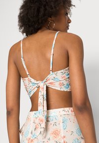 Top com padrão floral em bege claro, apresentando alças finas ajustáveis, um detalhe de laço nas costas e uma textura de tecido suave.
