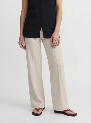 Person iført beige vidde bukser, sort ærmeløs vest med knapper og sorte sandaler med røde remme, stående mod ensfarvet baggrund.