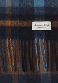 Wollschal in blau, braun und schwarz kariert mit Fransen. Das Etikett besagt: "Johnstons of Elgin, Made in Scotland." Weicher Textur, klassisches Design.