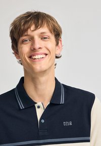 Marinblå och krämfärgad polo med tre knappar, horisontella ränder och texten "POLO CLUB" i vitt, tillverkad av en texturerad bomull.