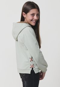 Sudadera verde claro con forro de estampado de leopardo, detalles de cordones laterales con acentos rosas y puños acanalados. Textura de tela suave.