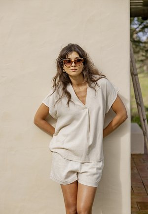 Žena v béžovom tričku s výstrihom do V a kraťasoch, stojaca s rukami za chrbtom, má na sebe slnečné okuliare s korytnačinovým vzorom a zlaté náušnice.