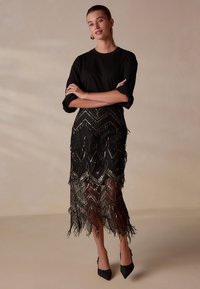 Black top with rolled sleeves, paired with a long black fringed skirt featuring a zigzag pattern and metallic accents. Black pointed heels.
