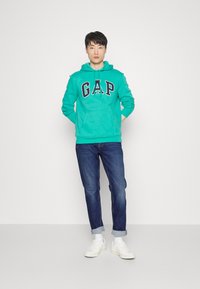 Felpa turchese con logo "GAP", jeans blu e sneakers bianche. La felpa ha un cappuccio con cordino e una tasca frontale. Vestibilità casual.