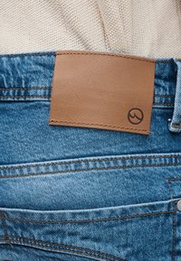 Μπλε τζιν παντελόνια με ραμμένη καφέ δερμάτινη ετικέτα στη μέση, που φέρει ένα μικρό μαύρο κυκλικό λογότυπο στη γωνία.