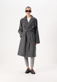 Siv plašč iz volne z obrečnim pasom, z zareznim ovratnikom in širokimi rokavi, nosi čez belo majico in sive kavbojke.