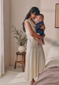 Kremowa dzianinowa kamizelka nałożona na zwiewną beżową spódnicę. Dziecko w kraciastym body, boso, trzymane na jasnym tle z rośliną i stołkiem.