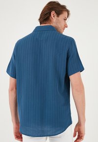 Cămașă cu mânecă scurtă dintr-un material texturat de culoare albastră închisă, cu dungi verticale subtile, având un guler clasic și un tiv curbat la spate.