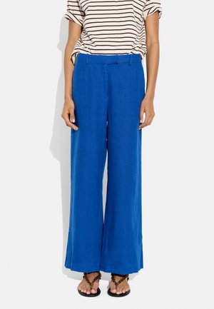 Kobieta w szerokich spodniach w kolorze kobaltowym, połączonych z białym i czarnym paskowanym wciągniętym do środka t-shirtem oraz brązowymi plecionymi sandałami.