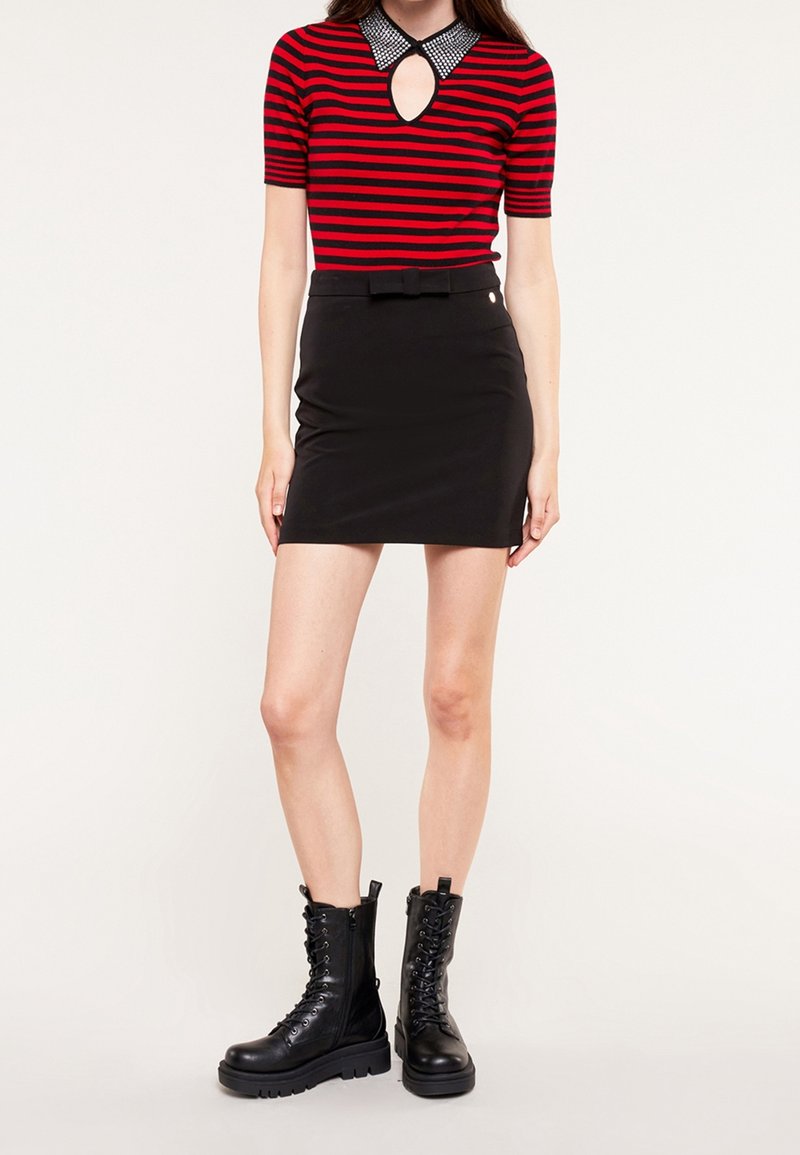Femme portant un top manches courtes rayé rouge et noir avec un décolleté en forme de goutte, une mini-jupe noire avec ceinture à nœud, et des bottes de combat noires à lacets.