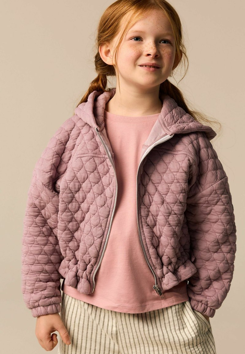 Junges Mädchen mit roten Haaren in Zöpfen, das eine gesteppte mauvefarbene Jacke über einem rosa Hemd und gestreiften hellen Hosen trägt und leicht lächelt.