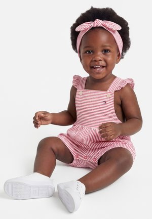 Šypsantis mažylis su afro šukuosena, vilkintis rožinės ir baltos spalvos dryžuotą kombinezoną, derančią galvos juostą ir baltus sportbačius, sėdintis prie balto fono.