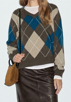 Donna che indossa un maglione a rombi marrone, blu e beige sopra una camicia bianca, gonna in pelle nera e porta una borsa a tracolla in camoscio color cuoio.