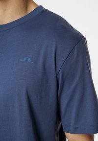 Sininen lyhythihainen t-paita, joka on valmistettu pehmeästä materiaalista, pyöreällä kaula-aukolla ja pienellä vaaleansinisellä logolla vasemmassa rinnassa.