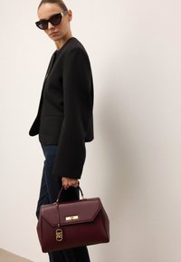 Bordowa skórzana torebka o wyraźnym kształcie, ze złotymi akcentami i odpinanym metkiem, noszona przez modelkę w czarnym żakiecie i dżinsach.