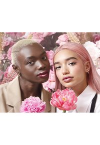 Deux individus posent de près, chacun tenant une grande pivoine rose. L'un a les cheveux blonds courts et porte un blazer beige ; l'autre a de longs cheveux roses et une chemise blanche.