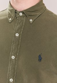 Camicia verde oliva con bottoni in tessuto strutturato, con colletto sagomato e logo nero ricamato sul petto sinistro. Bottoni bianchi.