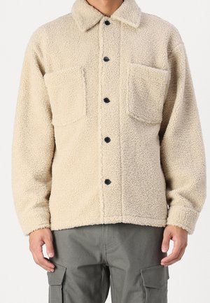 Personne portant une veste en polaire sherpa beige avec des boutons noirs et deux poches poitrine, associée à un pantalon cargo gris.