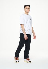 Camiseta blanca de manga corta con un logo negro, combinada con jeans negros y zapatillas multicolores con cordones estampados.