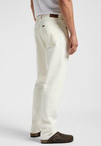 Jeans in denim bianco con taglio dritto, cuciture visibili e una patch in pelle marrone sulla vita. Indossati con zoccoli marroni.