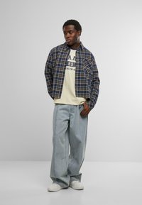 Fiatal férfi áll, kezét a zsebében tartva, kék kockás inget visel egy bézs grafikás póló felett, laza világos farmernadrágot és fehér sportcipőt.