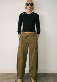 Maglione corto nero con dettagli a bottoni sulle spalle, abbinato a pantaloni cargo verde oliva con tasche laterali e orli risvoltati.