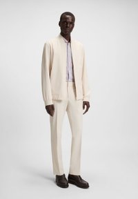 Chaqueta bomber crema sobre una camisa estampada, combinada con pantalones ajustados crema y zapatos marrón oscuro. Tejido suave con un diseño estructurado.