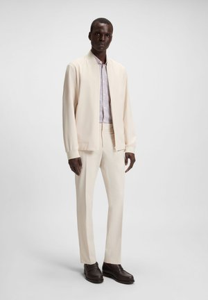Cream bomber jacket over a patterned shirt, paired with fitted cream trousers and dark brown shoes. Smooth fabric with a structured design.