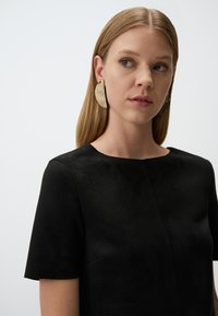 Zwarte suède top met korte mouwen en een ronde halslijn, met subtiele naaddetails. Grote metalen oorbellen zorgen voor contrast. Schone achtergrond.