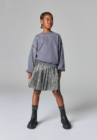 Sweatshirt gris avec des embellissements argentés, jupe plissée noire superposée, et bottines noires avec des chaussettes texturées. Tenue moderne et décontractée.
