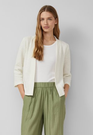 Kobieta o długich blond włosach nosi białą teksturowaną otwartą kurtkę, białą koszulkę i luźne oliwkowo-zielone spodnie, stoi na szarym tle.