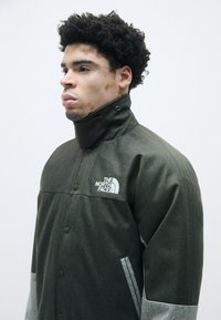 Sötétzöld halszálka mintájú zakó magas gallérral, patent gombokkal és szürke részletekkel, az elülső bal mellen a The North Face logójával.