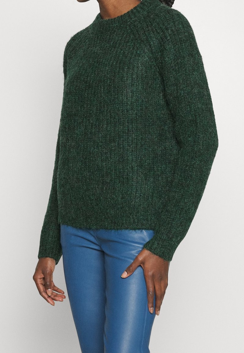 Pull à manches longues en tricot vert foncé porté avec un pantalon en cuir bleu vif moulant sur une personne debout.