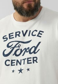 Behaarde man in een wit T-shirt met "SERVICE Ford CENTER" en drie sterren in donkerblauw op de voorkant.