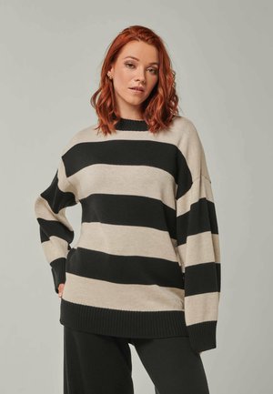 Frau mit schulterlangem roten Haar, die einen locker sitzenden schwarzen und beigen, horizontal gestreiften Pullover und schwarze Hosen vor einem einfarbigen Hintergrund trägt.