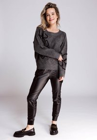 Grauer Strickpullover mit schwarzer Blumenstickerei, kombiniert mit glänzenden schwarzen Jogginghosen aus Kunstleder und schwarzen Loafers mit goldenen Akzenten.