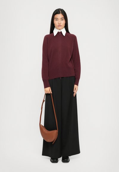 Mulher com cabelo preto liso, vestindo um suéter burdeos com fecho, camisa branca com colarinho, calças largas pretas e sapatos pretos, segurando uma bolsa castanha.