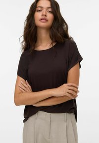 Haut marron à manches courtes en tissu doux, avec un col rond et une coupe décontractée. Le mannequin pose les bras croisés, portant un pantalon de couleur claire.