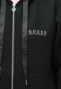 Μαύρο φούτερ με φερμουάρ και ασημένιες λεπτομέρειες, με λογότυπο που γράφει "GUESS LOS ANGELES", κορδόνια από σατέν κορδέλα και ριμπ υφή.