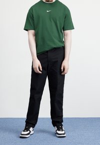 Green cotton T-shirt with short sleeves and Nike logo, paired with black pants and black-and-white sneakers, standing on blue carpet.