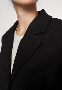 Blazer nero in lana con revers a vista, tessuto texturizzato e forma aderente, indossato su una camicia verde chiaro. Vista ravvicinata che mette in evidenza i dettagli del colletto.