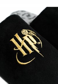 Black slippers with a soft texture, featuring a gold embroidered logo. The interior has a patterned gray fabric.