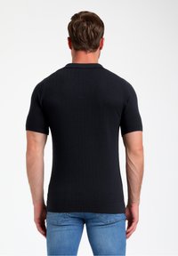 Melna īsām piedurknēm polo krekls ar ribotu tekstūru un pieguļošu dizainu, ar šauru apkakli un apakšmali. Valkāts ar gaiši ziliem džinsiem.