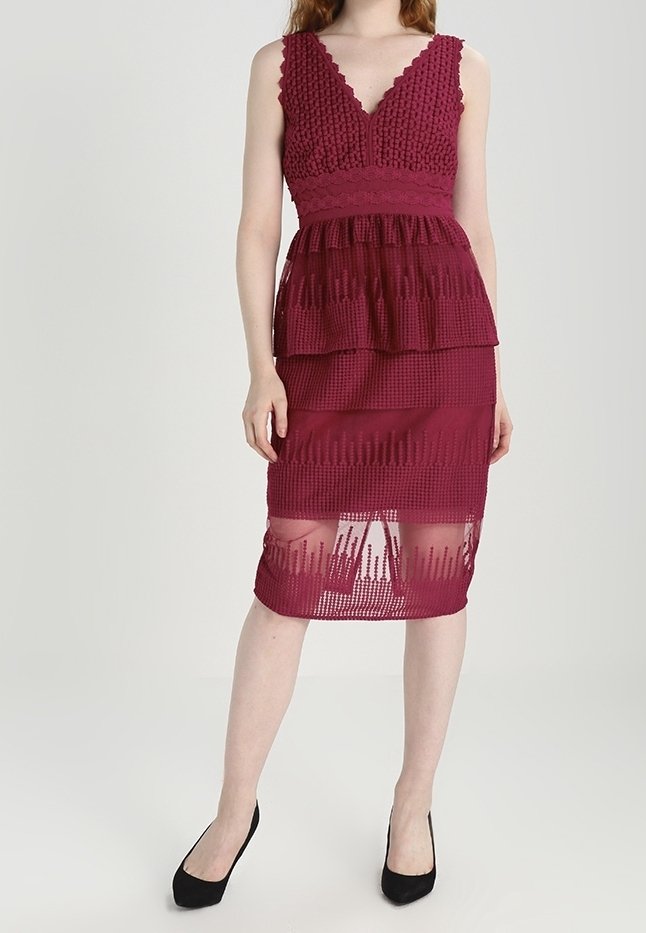 Femme portant une robe en dentelle bordeaux sans manches avec une jupe superposée et des motifs transparents, associée à des escarpins noirs à talons pointus, se tenant devant un fond uni.