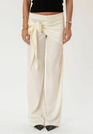 Žena oblečená v krémových širokých nohaviciach s detailom viazaným na mašľu v páse, čiernych špicatých topánkach a čiernom krátkom topi.