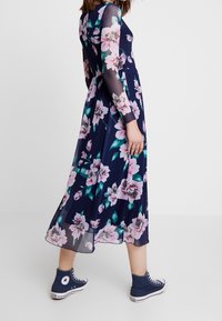 Robe longue fleurie bleu marine avec motifs floraux roses et verts ; manches longues transparentes ; associée à des baskets montantes bleues.