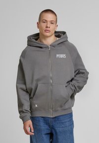 Šedá zipová mikina vyrobená z mäkkého materiálu, s veľkou kapucňou, prednými vreckami a vyšívaným logom "PEQUS" na ľavej hrudi.