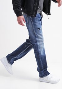Jeans i denim i en medel till mörkblå tvätt med en sliten reva på låret. Kombinerat med vita sportskor och en svart jacka.
