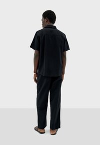 Chemise noire à manches courtes et pantalon assorti, fabriqués en tissu texturé, avec une coupe décontractée et un col ouvert, associés à des tongs noires.